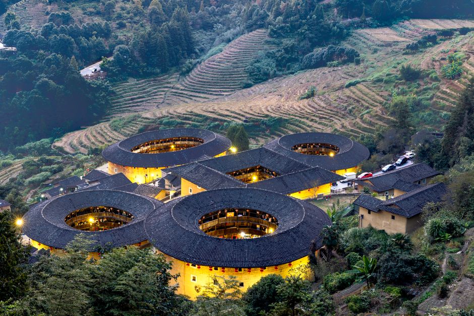 Scenic aerial view of illuminated Fujian Tulou amidst terraced fields in an autumn landscape.